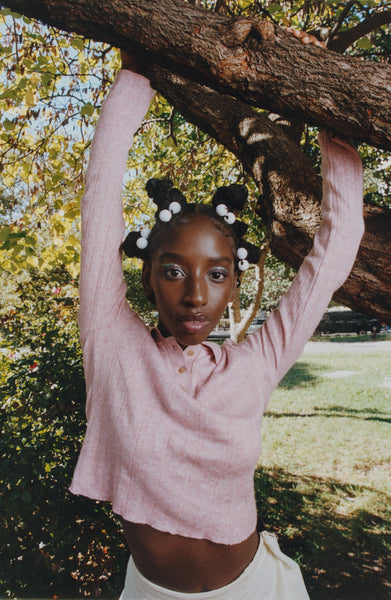 A Black woman wearing a pink shirt and glittery eye makeup grabs onto a tree holding her hands above her head