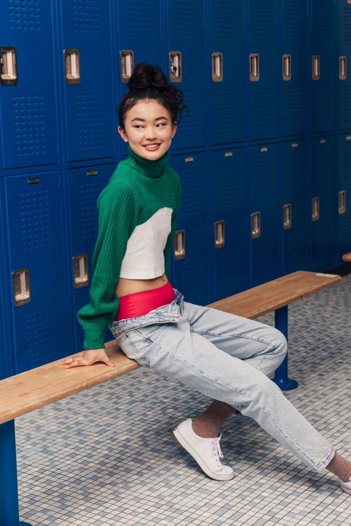 girl wearing jeans and green sweater sits on a locker room bench